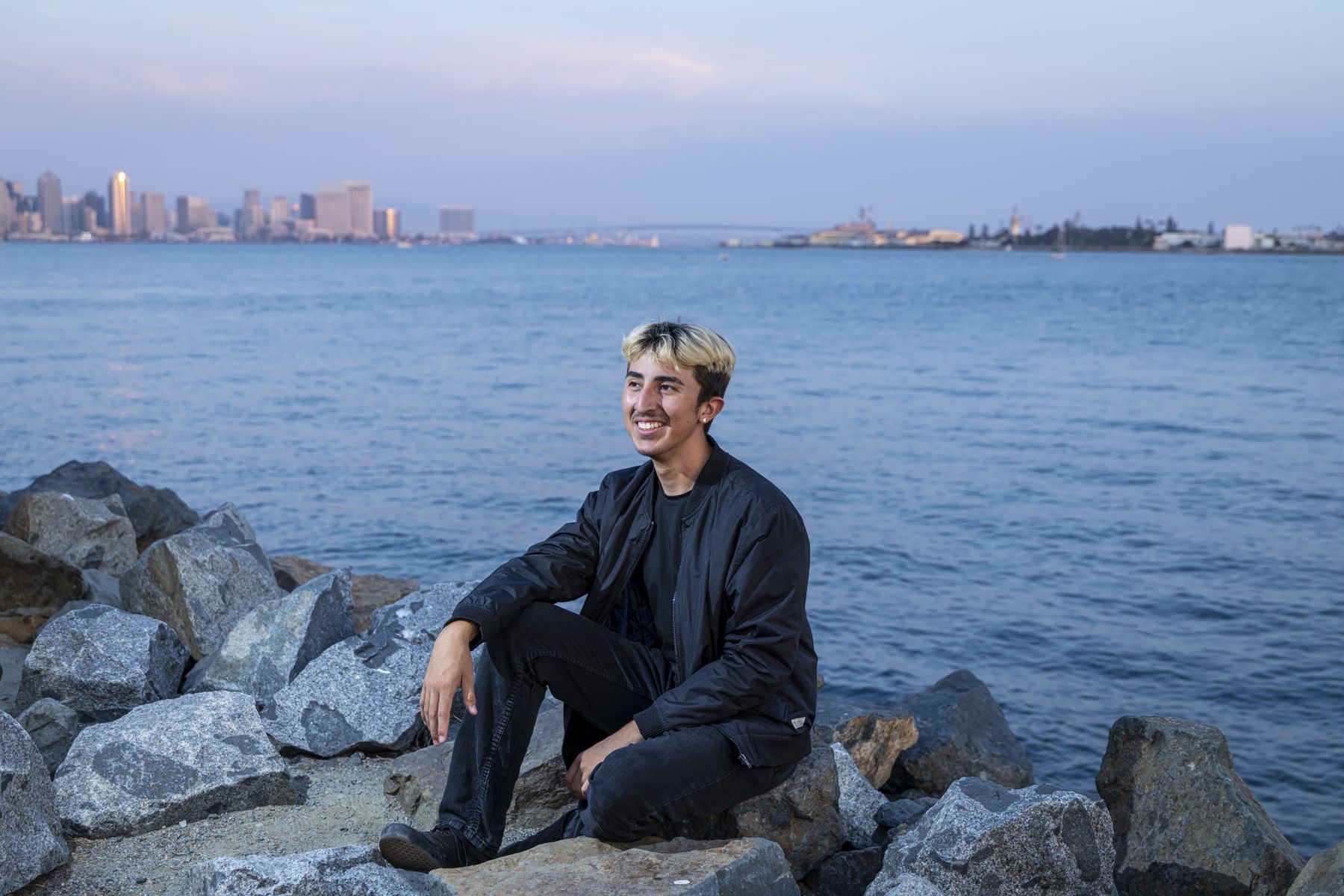 Esteban Lemus sitting on the rocks by San Diego Bay at dusk, downtown skyline behind him